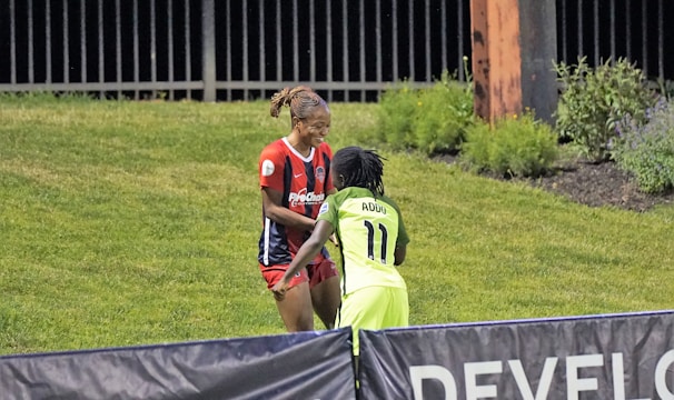 Two individuals are interacting on a grassy field. One is wearing a red and blue sports outfit with 'ProChain' visible, and the other is in a bright yellow uniform with the name 'Addo' and the number 11. They appear to be playing a sport, possibly soccer, as they are near a marked boundary. A fence and some vegetation are visible in the background.